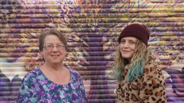 Two women standing in front of a shutter painted with tree design